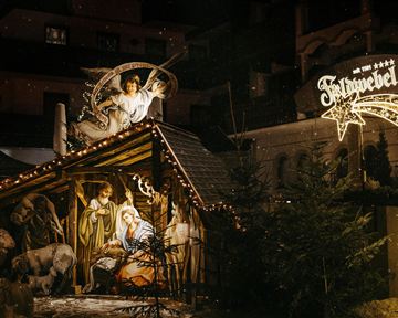 Eine festlich beleuchtete Weihnachtskrippe mit Figuren von Maria, Josef und dem Kind Jesus. Im Hintergrund ist ein geschmücktes Schild mit dem Namen des Ortes sichtbar.