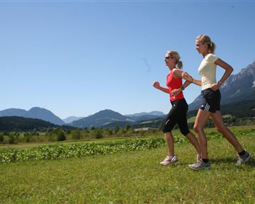 Zwei Frauen joggen auf einem grünen Feld mit Bergen im Hintergrund. Der Himmel ist klar und blau.