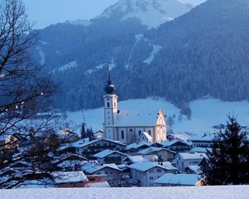 A picturesque winter landscape with a church in the foreground and snow-covered mountains in the background. The tranquil scene radiates peace and cold.