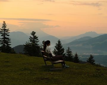 Eine Person sitzt alleine auf einer Bank und schaut auf eine malerische Berglandschaft während des Sonnenuntergangs. Die Szene ist von Bäumen umgeben und vermittelt eine ruhige, entspannende Atmosphäre.