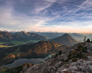 Eine beeindruckende Berglandschaft mit schneebedeckten Gipfeln und einem klaren See im Tal. Der Himmel ist von sanften Farben und Wolken durchzogen.