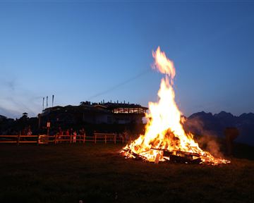 A large fire burns in a meadow at dusk. In the background, people and a building view are visible.