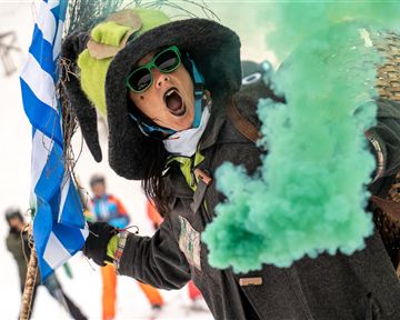Eine Person trägt einen großen Hut und Sonnenbrille und feuert eine grüne Rauchfackel ab. Im Hintergrund sind Menschen auf Ski zu sehen.
