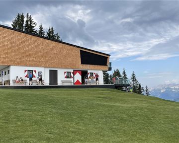 Ein modernes Gebäude mit einer schrägen Dachkonstruktion steht auf einer Wiese. Im Hintergrund sind Berge und ein bewölkter Himmel zu sehen.