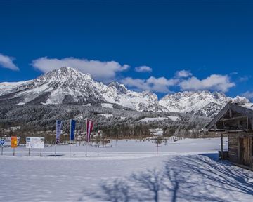 Eine schneebedeckte Landschaft mit majestätischen Bergen und einem klaren blauen Himmel. Im Vordergrund steht ein Holzgebäude und bunte Fahnen sind aufgestellt.