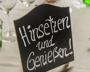 A sign with the inscription "Sit down and enjoy!" stands on a table. In the background, glasses and candles are visible.