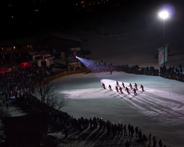 Eine Winterveranstaltung auf einer beleuchteten Eisfläche mit mehreren Schlittschuhläufern. Im Hintergrund sind eine große Menge Menschen und festliche Lichter zu sehen.