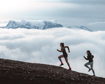 Zwei Frauen laufen einen hügeligen Weg hinauf, umgeben von Wolken. Im Hintergrund sind schneebedeckte Berge zu sehen.