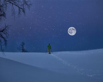 Ein skier steht in der verschneiten Landschaft unter dem Vollmond. Der Himmel ist dunkelblau und es schneit leicht.