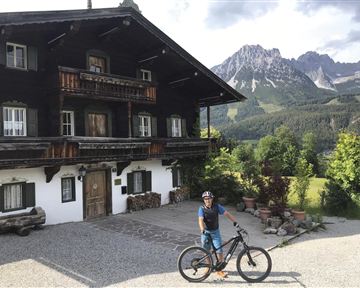 Ein Mountainbiker steht vor einem traditionellen Holzhaus in den Bergen. Im Hintergrund sind die Gipfel und die grüne Landschaft zu sehen.
