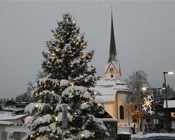 A festively decorated Christmas tree in front of a snow-covered church. The sky is gray and the surroundings feel wintry and cozy.