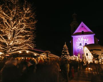 A festive Christmas market at night with brightly lit stalls. In the background, there is a church with purple lighting and a decorated Christmas tree.