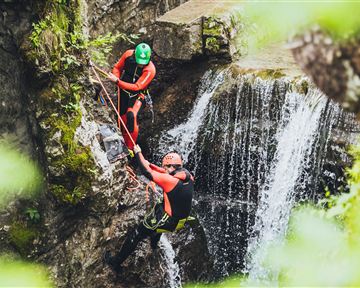 Two people in wetsuits climbing a rock wall. In the background, a waterfall is visible.