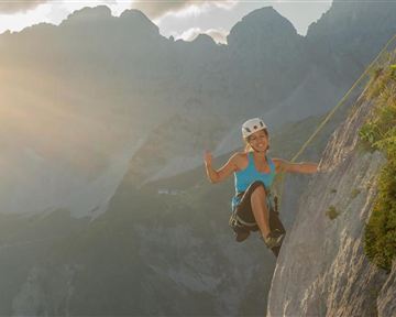 Eine Person klettert an einer steilen Felswand in den Bergen. Die Szene wird von Sonnenlicht erleuchtet und zeigt die Freude des Kletterns.