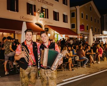 Zwei Musiker stehen lächelnd auf der Straße, einer mit einer Trompete und der andere mit einem Akkordeon. Im Hintergrund sind viele Menschen in einem beleuchteten Restaurant und auf der Terrasse zu sehen.