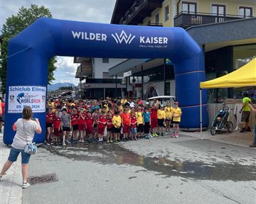 Eine Gruppe von Kindern steht unter einem großen blauen Startbogen, der den Schriftzug "Wilder Kaiser" trägt. Im Hintergrund sind Gebäude und eine gelbe Zeltstation sichtbar.