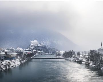 Eine verschneite Stadt am Ufer eines Flusses. Im Hintergrund sind Berge und grauer Himmel zu sehen.