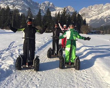 Four people are riding Segways through a snowy landscape. In the background, mountains and forests are visible.