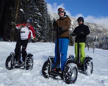 Drei Personen fahren auf funktionsfähigen Segways durch schneebedeckte Landschaft. Im Hintergrund sind Bäume und Berge sichtbar, und der Himmel ist klar.