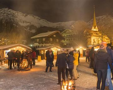 Ein winterlicher Weihnachtsmarkt bei Nacht mit festlich beleuchteten Ständen. Menschen genießen die Atmosphäre in einer schneebedeckten Umgebung, umgeben von Bergen.