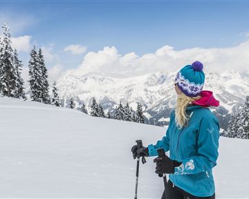 Eine Person steht im Schnee und blickt auf die verschneiten Berge. Sie trägt eine warme Jacke und eine Mütze, umgeben von einer winterlichen Landschaft.