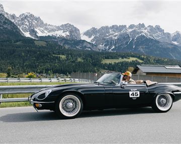 A black convertible drives along a winding road with impressive mountains in the background. The driver is wearing a hat and enjoying the ride in nature.