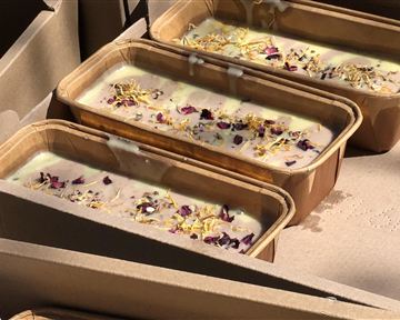 Drei Formen mit Seife, garniert mit getrockneten Blüten, liegen in einem Karton. Die Seife hat eine cremige Textur und eine helle Farbe.