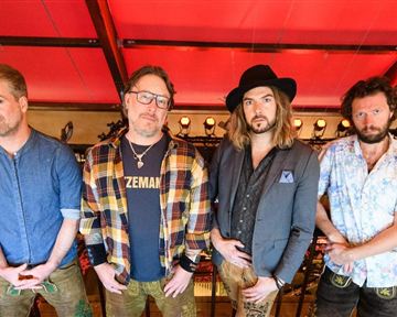 Eine Gruppe von vier Männern steht auf einer Tribüne. Sie tragen verschiedene Outfits und posieren vor einem roten Hintergrund.