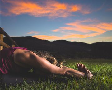 Eine Person macht Yoga im Gras mit einer wunderschönen Berglandschaft im Hintergrund. Der Himmel ist bei Sonnenuntergang in sanfte Farben getaucht.