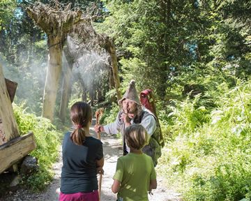 A hiker in the woods tells stories to children. The surroundings are green and idyllic, with many plants and trees.