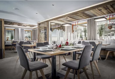 Ein modernes Restaurant mit eleganten Holztischen und stilvollen Stühlen. Große Fenster sorgen für viel Tageslicht und eine einladende Atmosphäre.