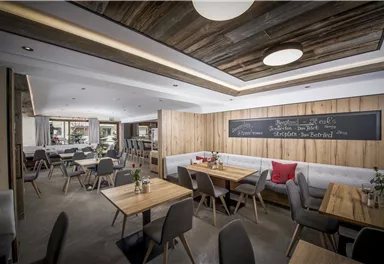 Ein modernes Restaurant mit Holzmöbeln und gemütlicher Atmosphäre. Tische sind ordentlich gedeckt und eine schwarze Tafel mit Schrift ist an der Wand zu sehen.