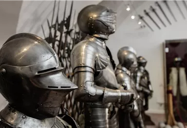 A series of medieval armors are displayed in a museum. In the background, various weapons can be seen.