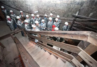 Eine Gruppe von Menschen in silbernen Anzügen und Helmen steht an einer Treppe in einem historischen Gebäude. Die Wände sind aus Stein und die Atmosphäre wirkt industriell und spannend.