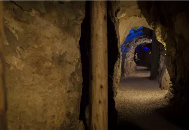 Ein unterirdischer Tunnel mit ausgehöhlten Wänden und sanfter Beleuchtung. Der Boden ist mit kleinen Steinen bedeckt und die Wände sind aus natürlichem Gestein.