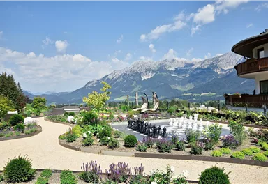 A beautiful garden landscape with colorful plants and a water basin. In the background, impressive mountains can be seen under a blue sky.