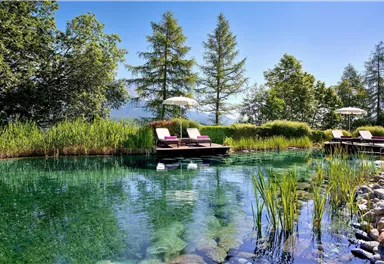 A calm, clear pond surrounded by lush greenery and tall trees. Relaxed lounge chairs provide a perfect spot to unwind.