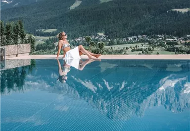 Eine Frau entspannt am Rand eines Schwimmbeckens mit Blick auf majestätische Berge. Das Wasser spiegelt die Umgebung und der Himmel ist klar und blau.
