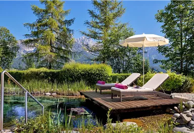 A relaxing outdoor area with sun loungers and a parasol. Surrounded by trees and a clear water basin, it offers a tranquil atmosphere.