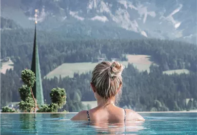 Eine Frau steht im Wasser eines Pools und blickt auf die Berge. Die Landschaft ist malerisch mit grünen Wiesen und einer Kirche im Hintergrund.