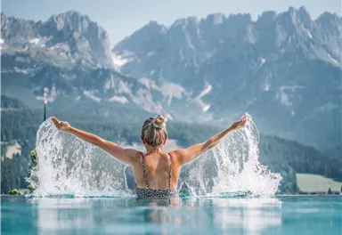 Eine Frau im Wasser eines Infinity-Pools genießt die Aussicht auf die Berge. Spritzendes Wasser umgibt sie und reflektiert die Schönheit der Natur.