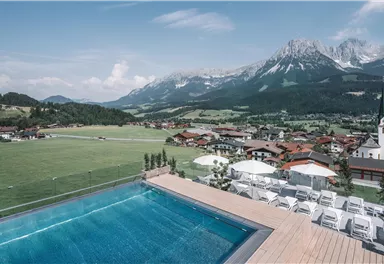 Ein atemberaubender Blick auf die Berge und ein Schwimmbad auf einer Terrasse. Im Hintergrund sind malerische Häuser und eine Kirche zu sehen.
