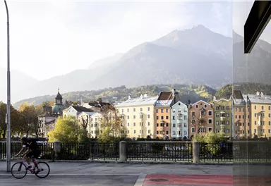 A picturesque cityscape with colorful buildings and the Alps in the background. A person is riding a bicycle along the road.