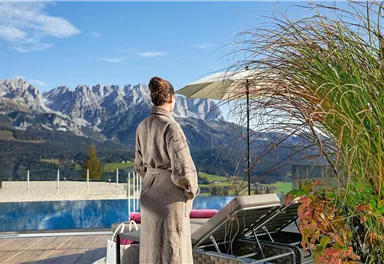 A person in a bathrobe stands at the edge of a pool and looks out at a mountainous landscape. The sky is clear and the surroundings feel relaxed and inviting.