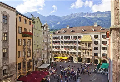 A bustling square in a city with historical buildings and mountains in the background. People are enjoying the atmosphere and the sun.