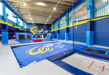 Eine Trampolinhalle mit blauen Matten und mehreren Trampolinen. Die Wände sind in Blau und Gelb gestrichenen, und es gibt viel Platz zum Springen und Spielen.