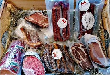 A rustic wooden basket with various types of meat and sausages. The products are arranged in transparent packaging and lie on hay.