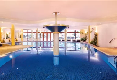 An elegant indoor pool with blue water and a modern water fountain. The surroundings are bright and inviting, with windows that let in natural light.