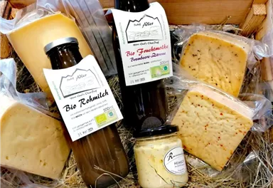 A rustic basket with various types of cheese and organic raw milk. The products are packaged in plastic and draped on hay.