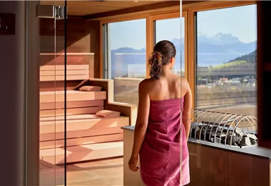 A woman in a towel looks out of a large window at the mountains. The room is modern and calm, with wooden and glass elements.